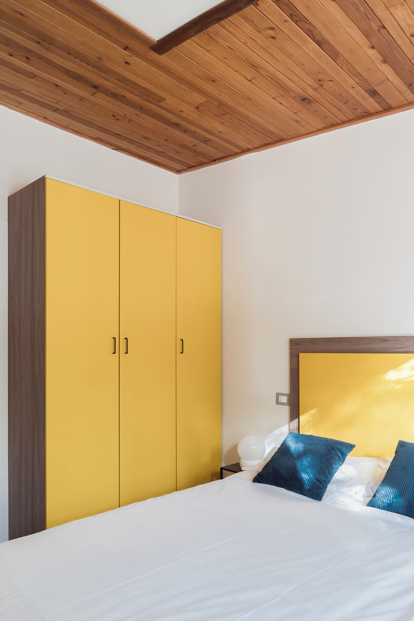 Une chambre avec un plafond en bois, une armoire jaune, un lit blanc avec des coussins bleus et un grand miroir au-dessus du lit.