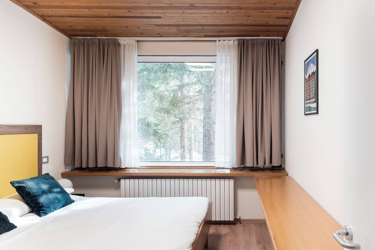 A bedroom with a bed, a window with curtains, a radiator, and a wooden bench.