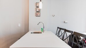 Une cuisine de taille réduite, dotée d’un plan de travail blanc, d’un évier en acier inoxydable et d’une seule chaise.