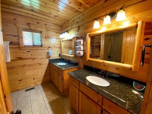 Une salle de bain de style rustique, avec des murs en bois, deux lavabos, un grand miroir et une fenêtre équipée de stores.