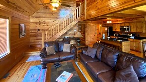 A cabin interior with a wooden interior, a sofa, a coffee table, a kitchen area, and a staircase. - Easy Drive Resort Cabin w/ Hot Tub, Mtn View, and Resort Pool Near National Park (Gatlinburg)