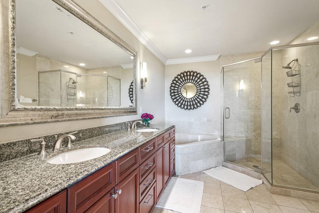 A modern bathroom with a granite countertop, two sinks, a large mirror, a round wall decoration, a glass shower enclosure, and a white bath tub.