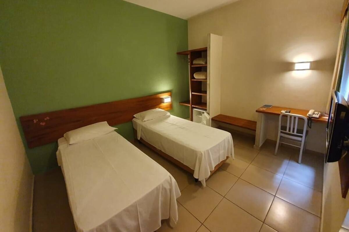 Ein Hotelzimmer mit zwei Einzelbetten, einem Schreibtisch mit Stuhl, einem Regal mit Handtüchern und einer Nachttischlampe.