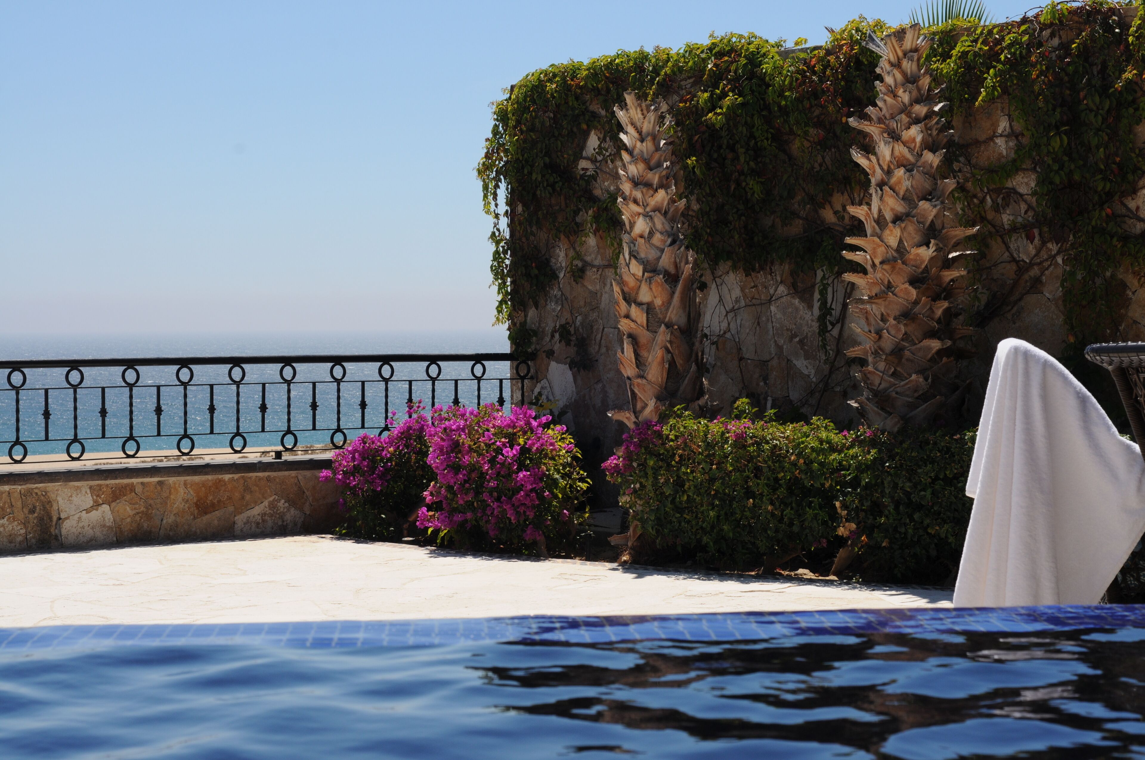 A pool area with a view of the sea, a white chair, and a stone wall with greenery.