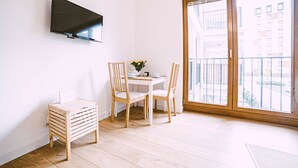 A modern room with a flat-screen TV mounted on the wall, a small wooden side table, a white dining table with two chairs, and a large window with a view of buildings. - Apartment Go at Rakowicka street  located just few minutes walk from city center (Kraków)
