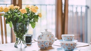 A table with a vase of yellow roses, a teapot, and matching cups on a wooden table in a room with large windows and curtains. - Apartment Go at Rakowicka street  located just few minutes walk from city center (Kraków)