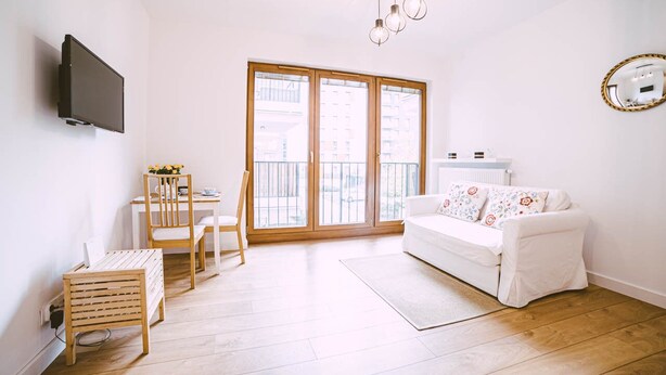 A modern living room with a white sofa, a dining table with chairs, a TV mounted on the wall, and a large window with a view of buildings. - Apartment Go at Rakowicka street  located just few minutes walk from city center (Kraków)