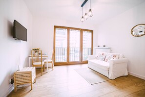 Un salon moderne avec un canapé blanc, une table à manger avec des chaises, un téléviseur fixé au mur et une grande fenêtre donnant sur des immeubles.