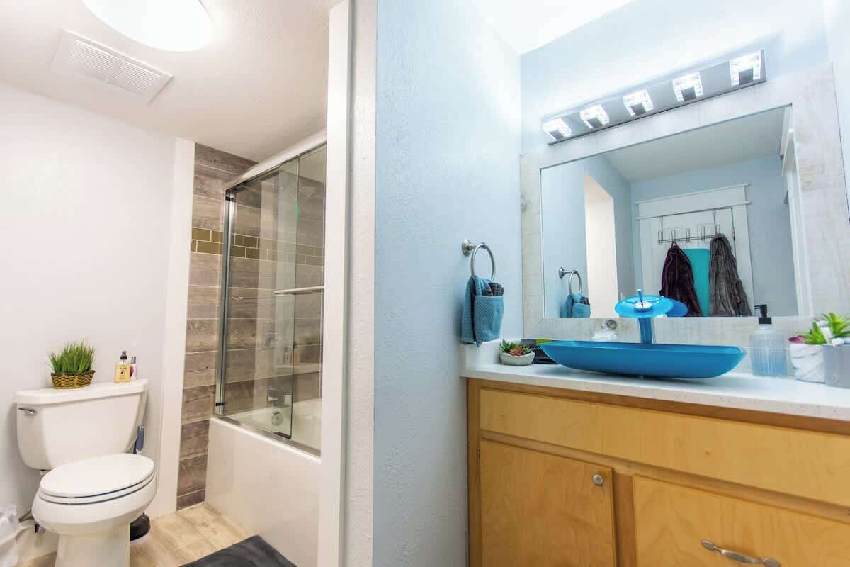 A modern bathroom with a glass shower enclosure, a white toilet, a blue sink with a large mirror, and wooden cabinets.