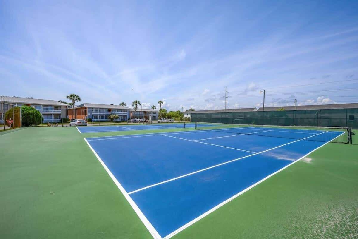 A tennis court with a green and blue surface, surrounded by buildings and palm trees.