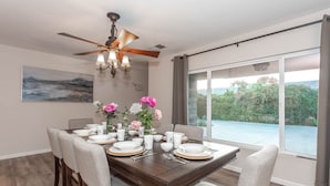 Une salle à manger avec une table en bois foncé, dressée pour six personnes, des chaises à l’assise rembourrée de couleur gris clair, un ventilateur de plafond et une grande fenêtre donnant sur un paysage verdoyant.