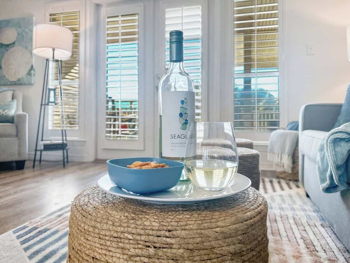 Una sala de estar con un taburete redondo tejido, un tazón azul con pasta, una botella de vino y una copa de vino blanco.