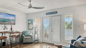 Una sala de estar con comedor, un ventilador de techo, un televisor y un letrero que dice 'BEACH HOUSE'.