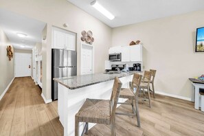 Een moderne keuken met een roestvrijstalen koelkast, witte kasten, een magnetron en een koffiezetapparaat. Er is een granieten aanrecht en rieten stoelen rond een klein eethoekje.