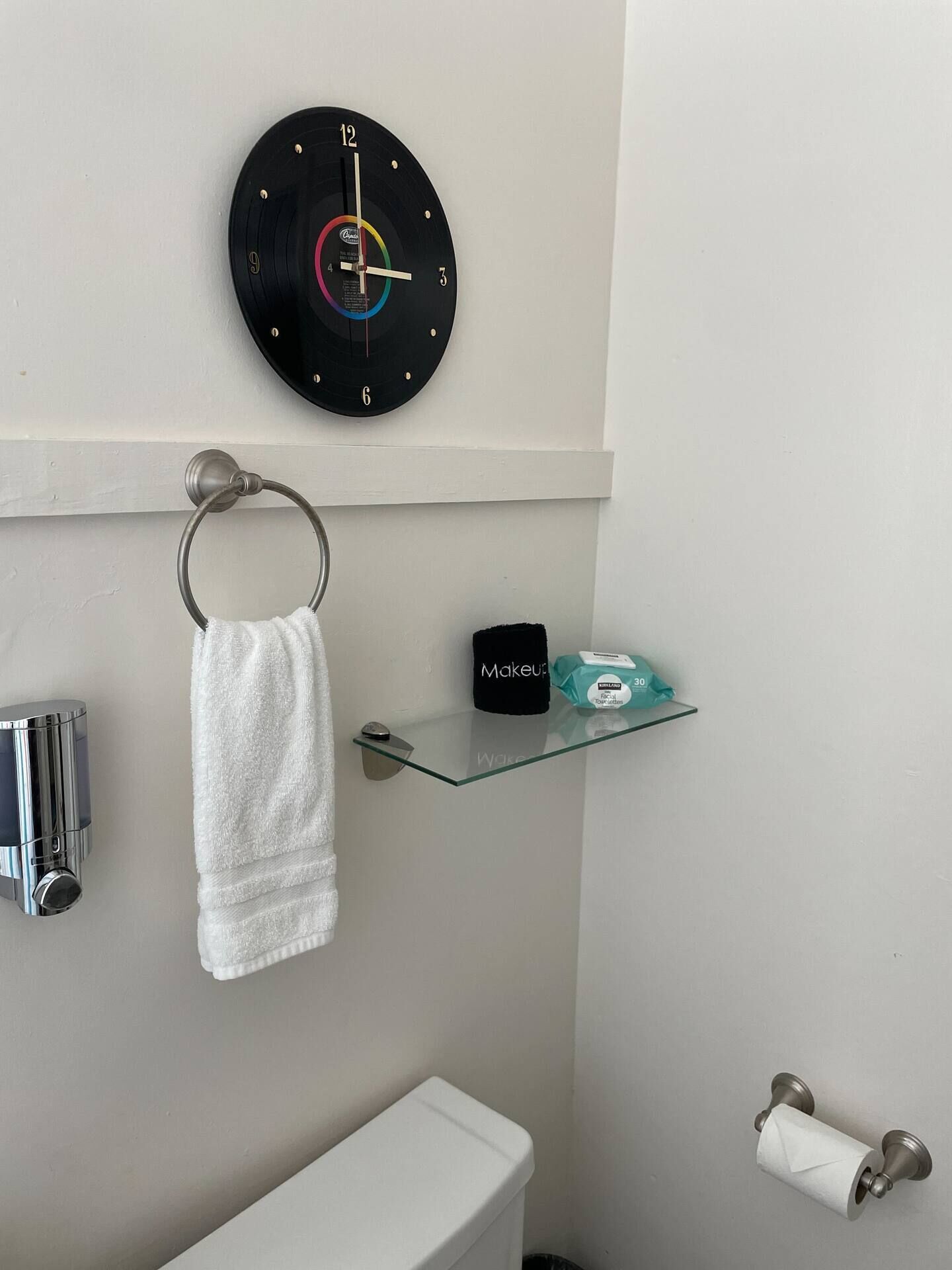 Uma casa de banho com um relógio de parede redondo, um suporte de toalha, uma prateleira de vidro com artigos de higiene e um porta-papel higiénico.