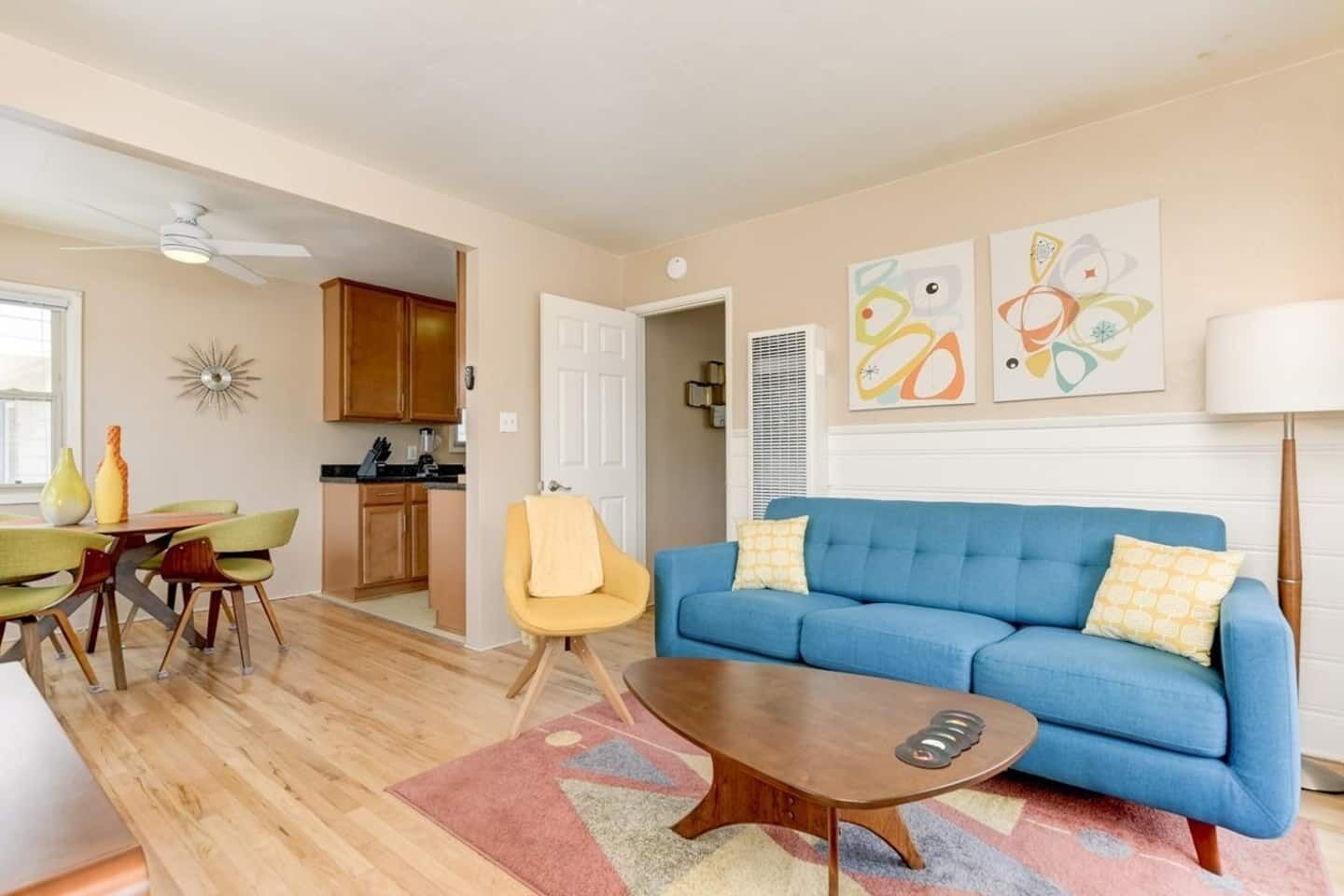 Ein modernes Wohnzimmer mit einem blauen Sofa, einem Holztisch und einem gelben Sessel. Es gibt einen Essbereich mit Tisch und Stühlen, und im Hintergrund eine Küche.