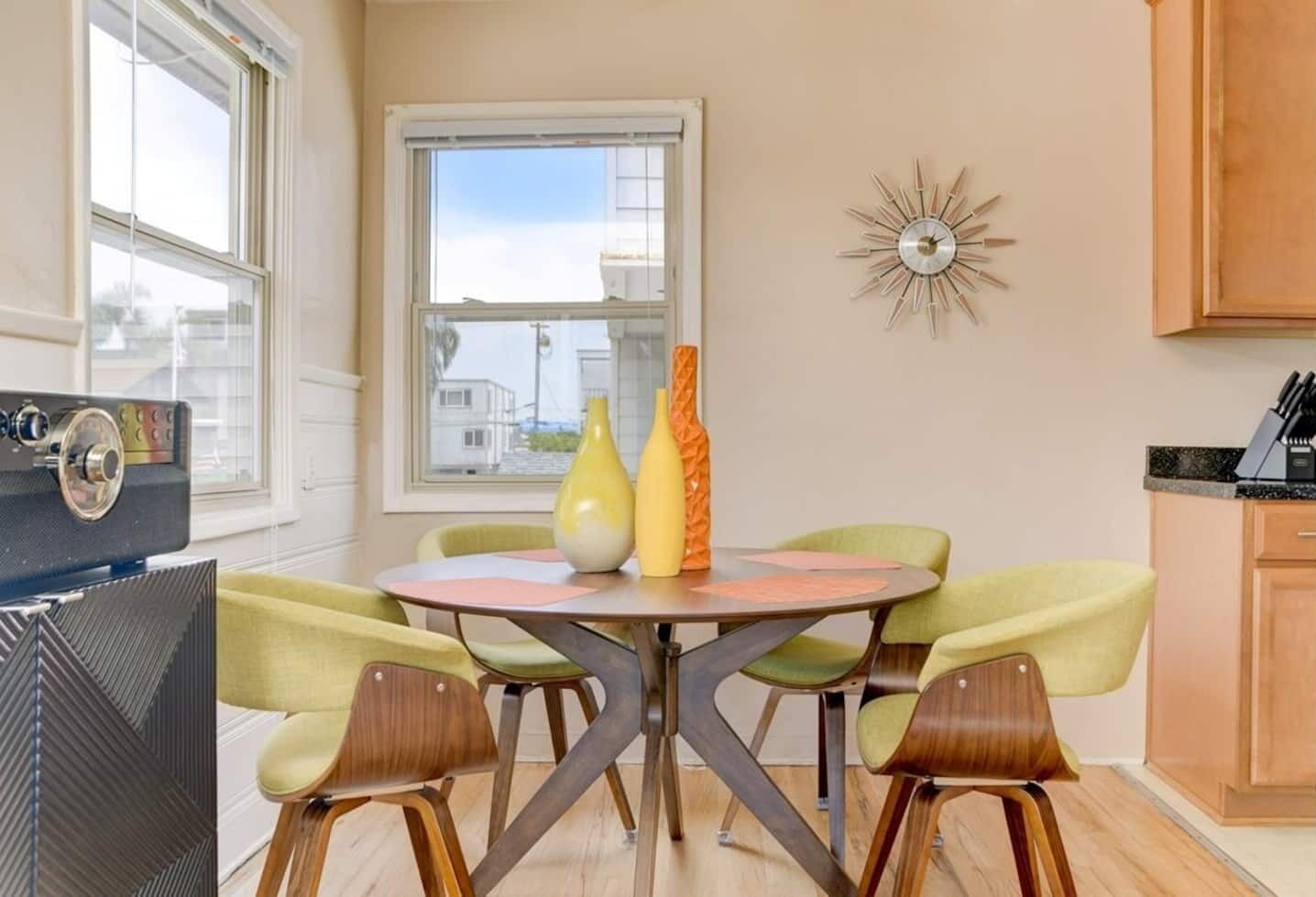 A dining area with a round table, four chairs, and two vases. There is a window with a view of buildings outside.