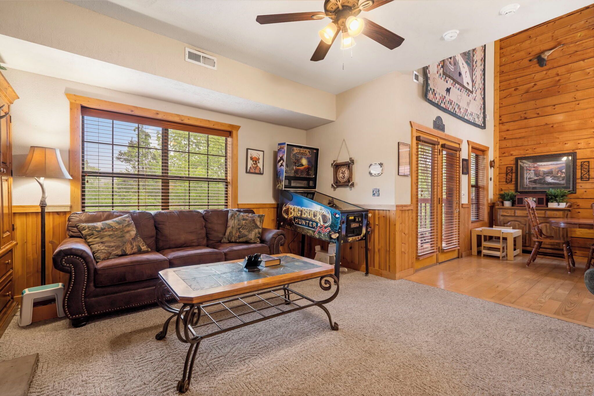 A cozy living room with a brown leather couch, a coffee table, and a ceiling fan. There is a window with blinds, a fireplace, and a television mounted on the wall.