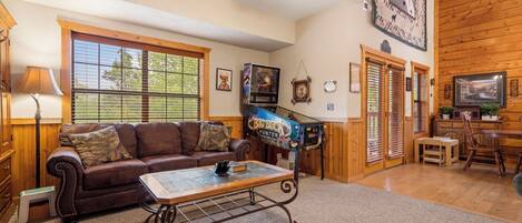 A cozy living room with a brown leather couch, a coffee table, and a ceiling fan. There is a window with blinds, a fireplace, and a television mounted on the wall.