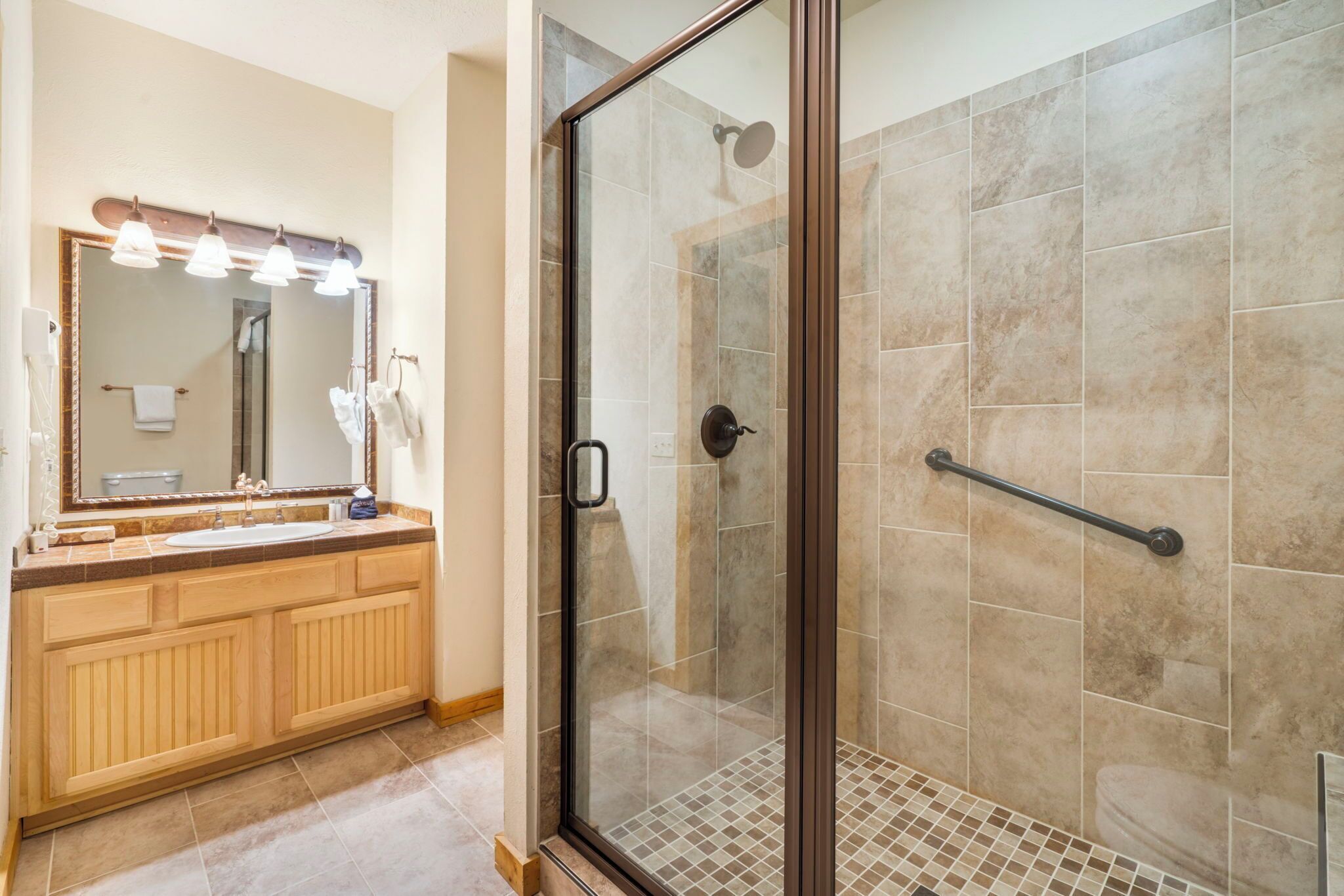 A bathroom with a glass-enclosed shower, a vanity with a mirror and lights, and a tiled floor.