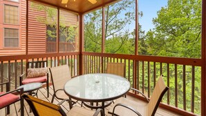 Un espacio al aire libre techado con una mesa redonda de vidrio y sillas, barandillas de madera y vistas a árboles.