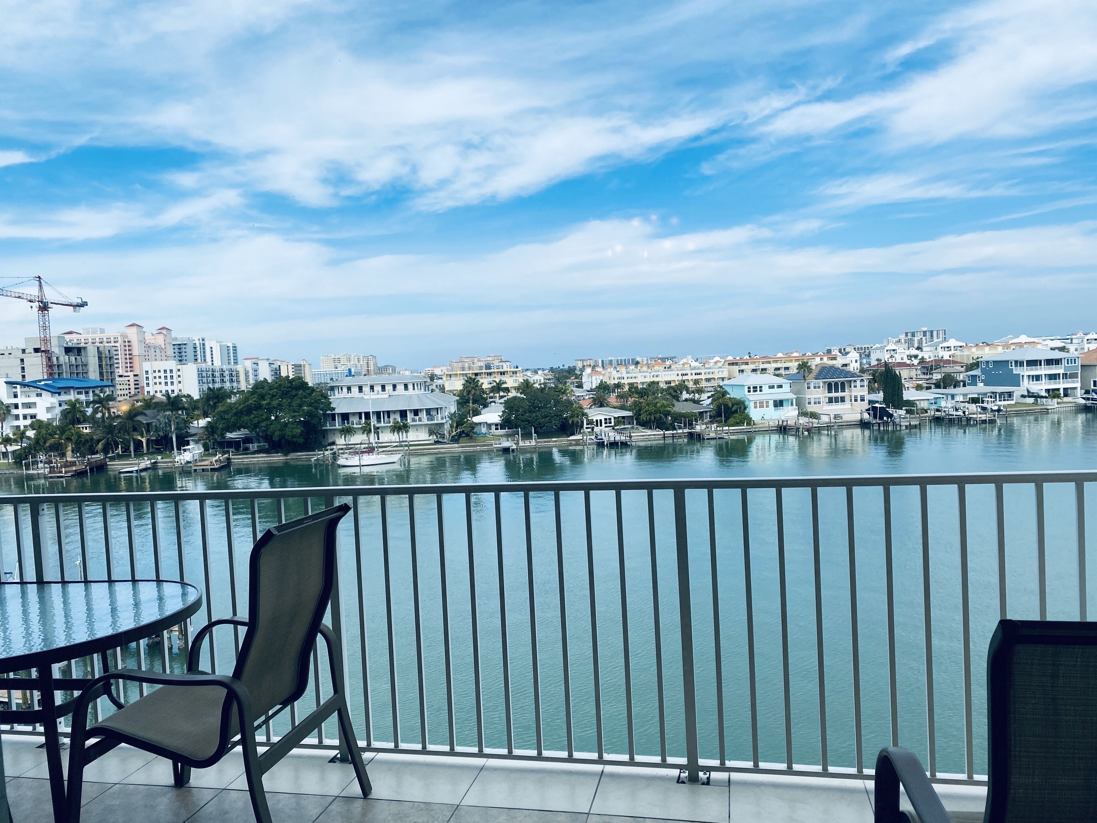 Un balcon avec une table et des chaises donnant sur une rivière paisible, avec des bâtiments et une grue en arrière-plan.