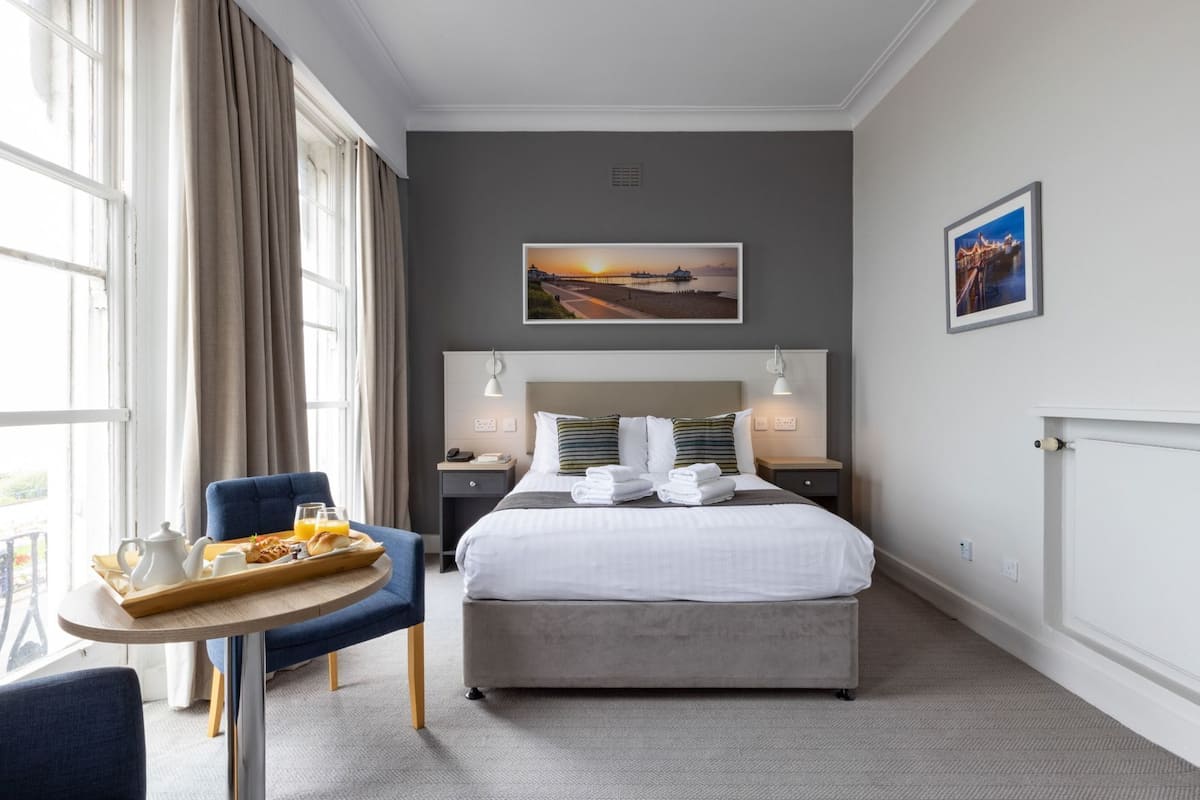 A hotel room with a bed, bedside tables, a chair, a small table with breakfast items, and two wall-mounted lamps.