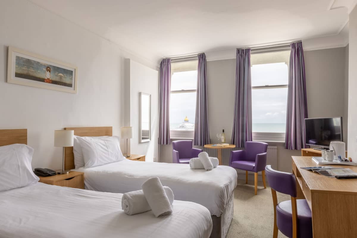A hotel room with two beds, a desk, a TV, and a view of the sea.