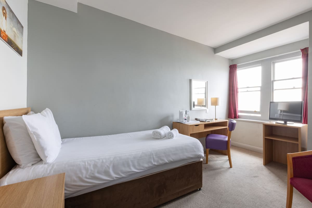 A hotel room with a single bed, a desk with a chair, a TV, and a window with curtains.