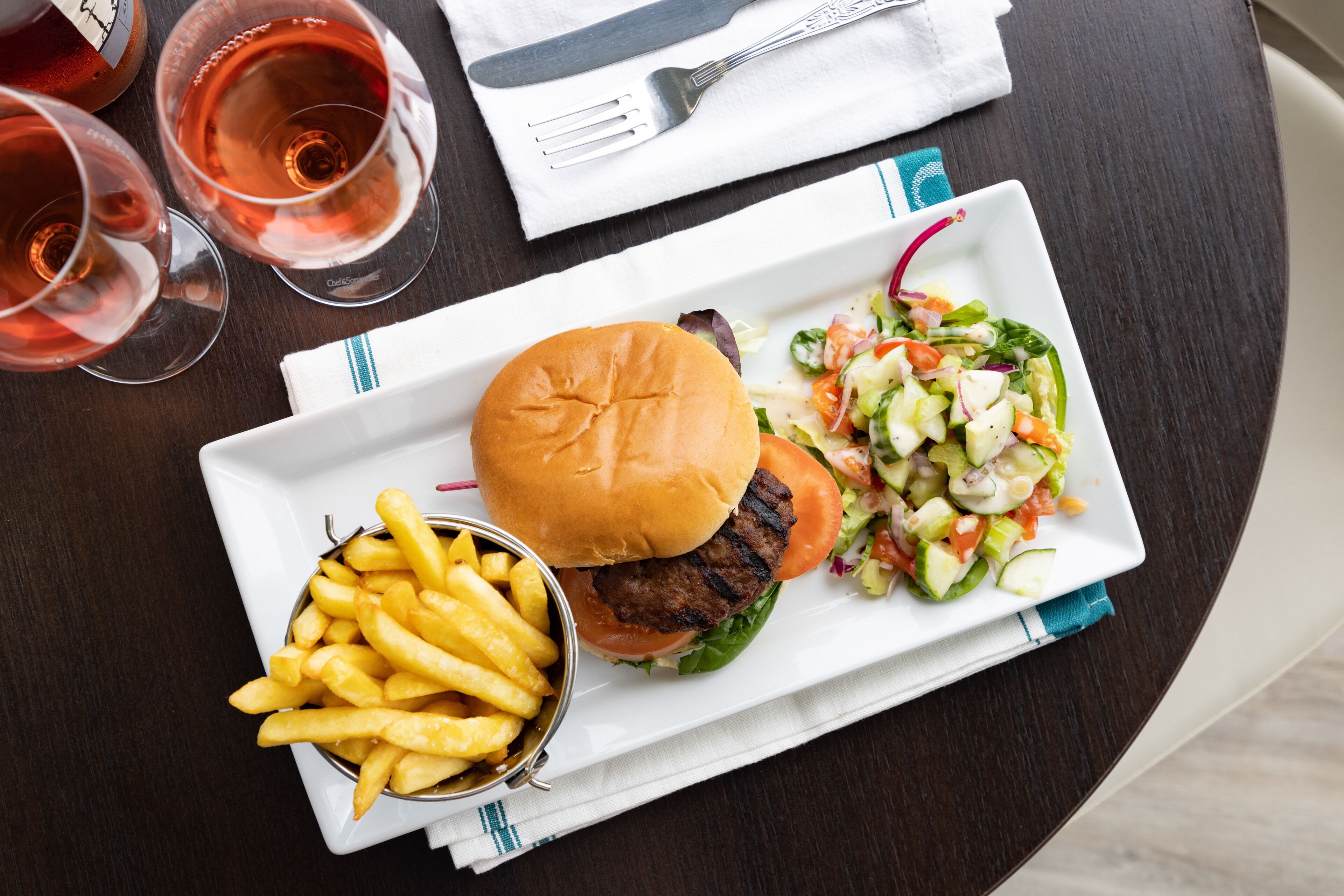 A meal consisting of a burger, fries, and a salad on a white plate, with two glasses of rosé wine on the table.