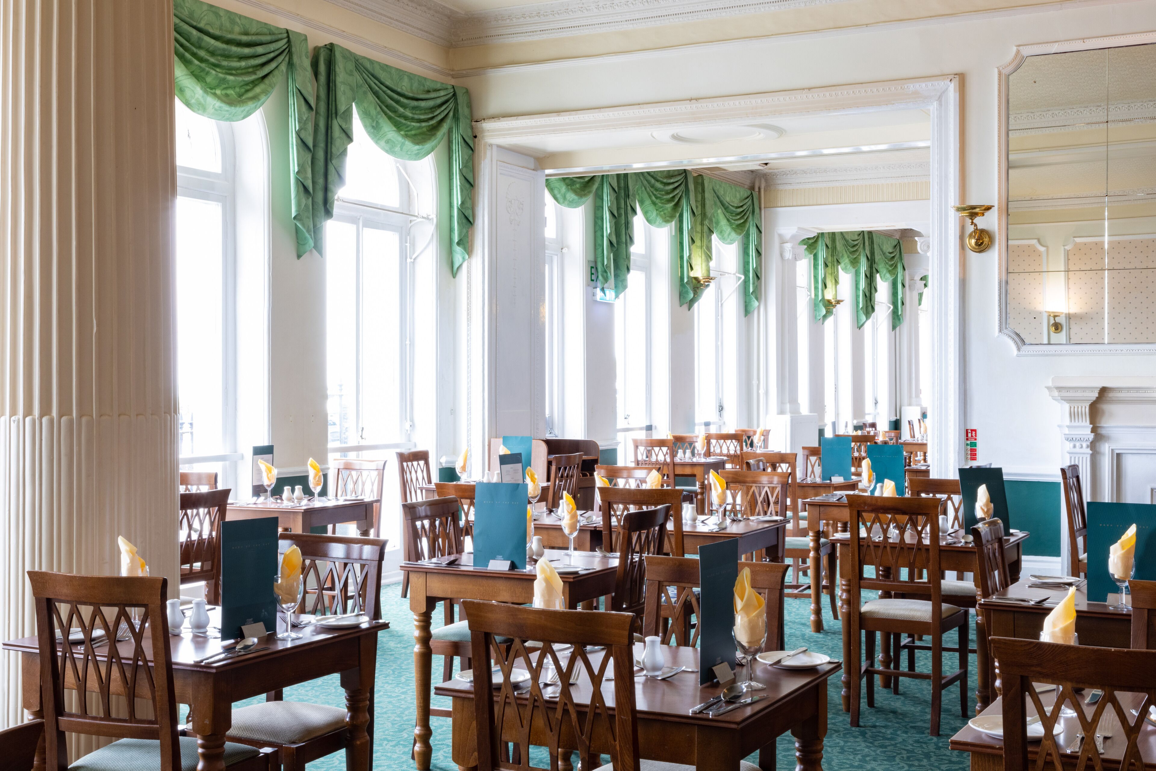 A dining area with multiple wooden tables and chairs, green drapes, and a chandelier.