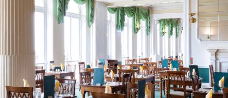 A dining area with multiple wooden tables and chairs, green drapes, and a chandelier.
