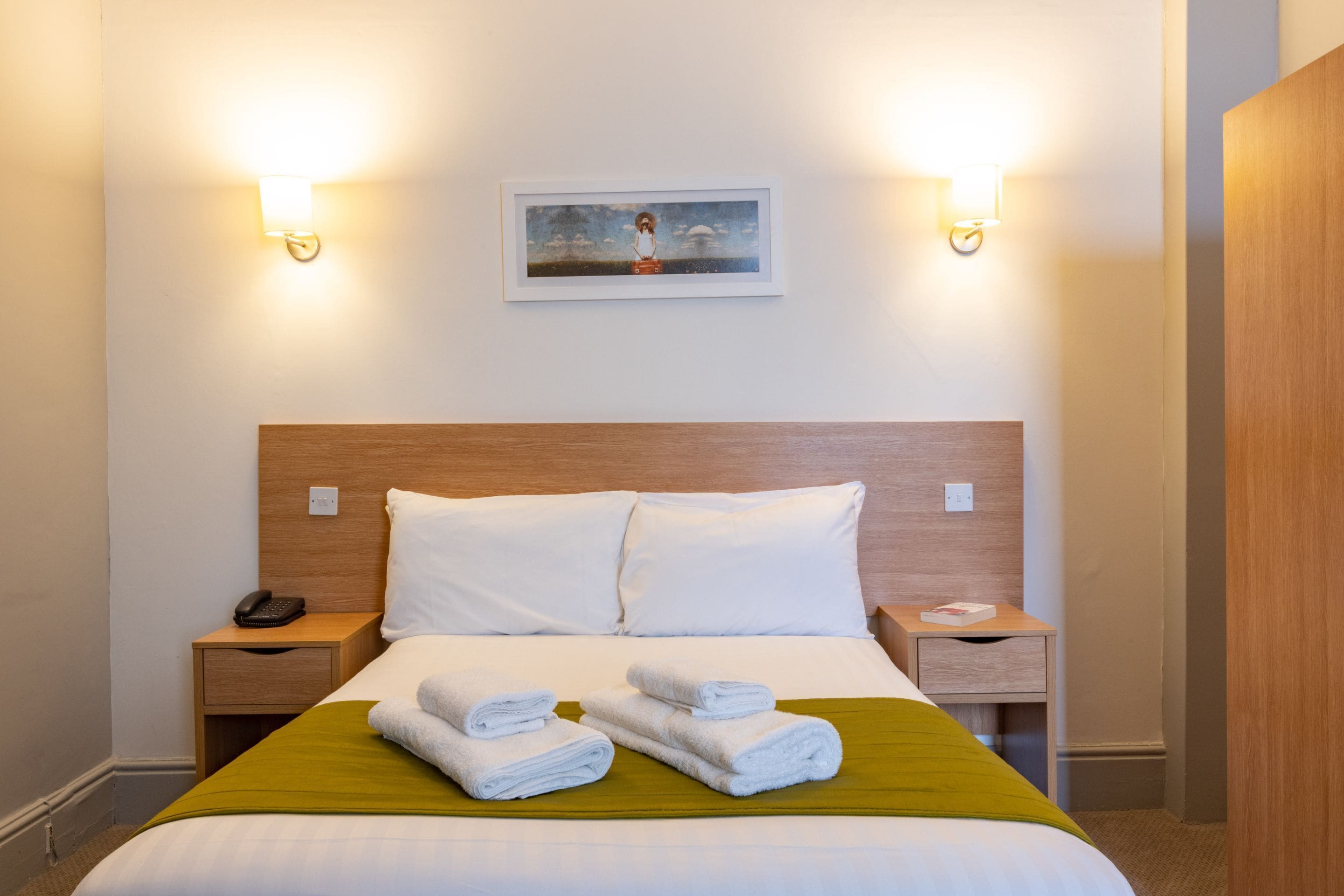 Una cama bien hecha con sábanas blancas y dos toallas enrolladas, un cabecero de madera y dos mesitas de noche con lámparas.
