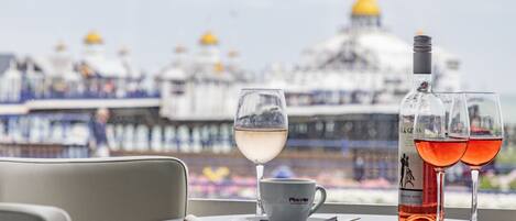 A table with a white cup and saucer, a bottle, and two glasses of wine, with a view of a ship and a building in the background.