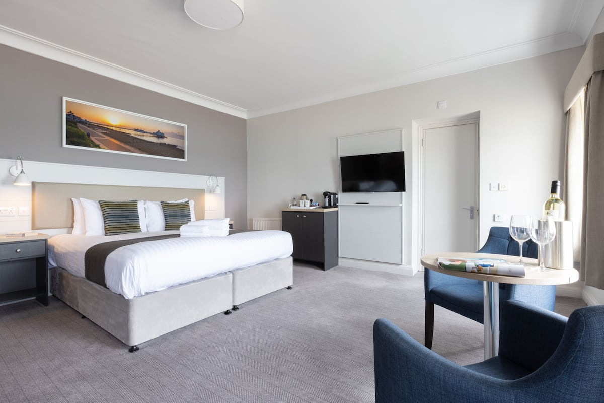 A modern hotel room with a large bed, a desk, a dining area, and a TV on the wall.