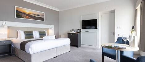 A modern hotel room with a large bed, a desk, a dining area, and a TV on the wall.