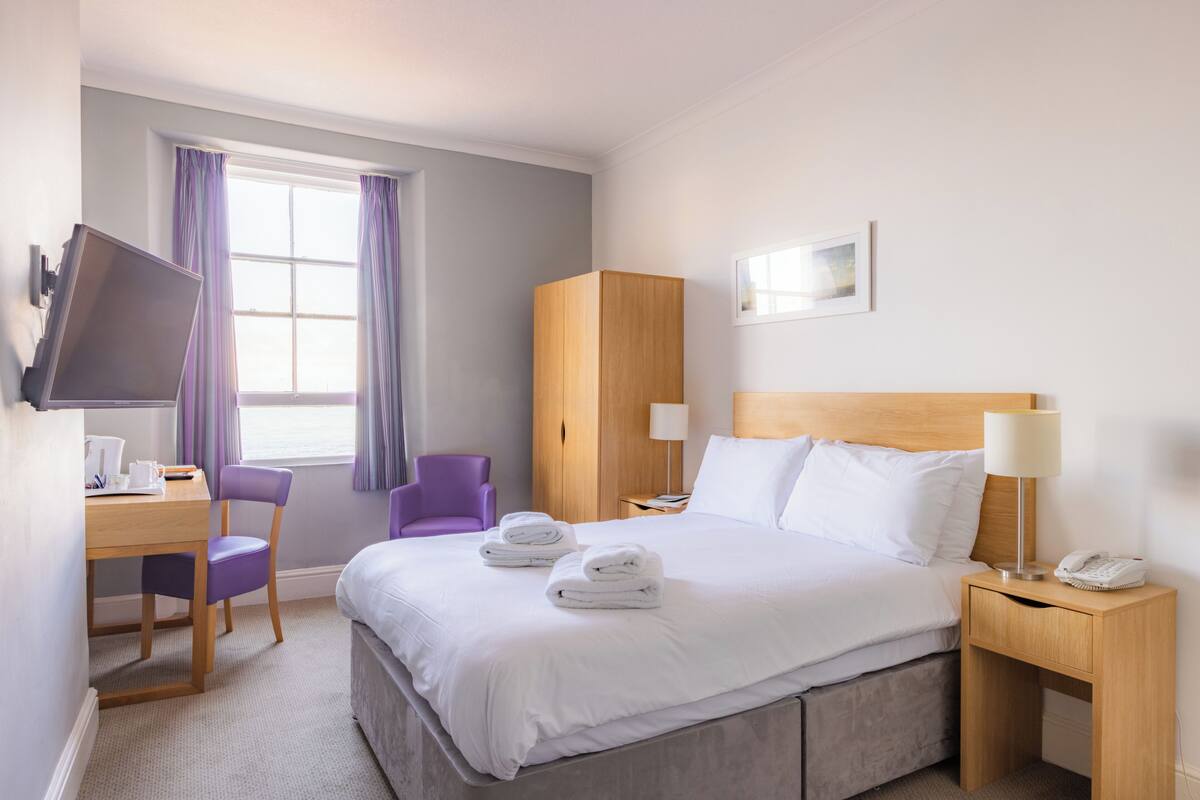 A hotel room with a bed, a desk with a chair, a wardrobe, a TV, and a window with curtains.