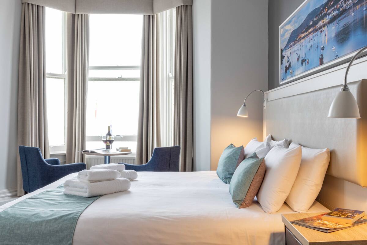 A hotel room with a neatly made bed, a side table with a lamp, a chair, and a view of the sea through large windows.
