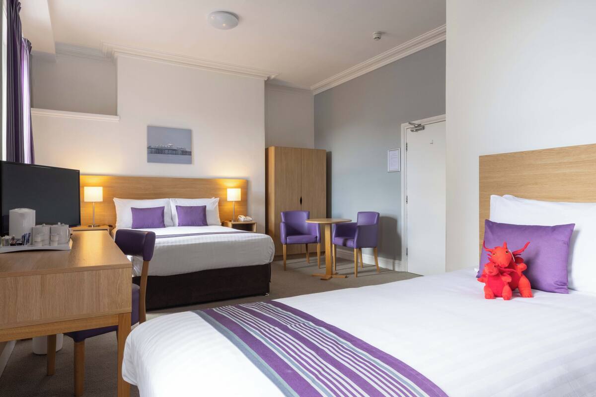 A hotel room with a bed, a desk with a lamp, a chair, a small table, and a television.