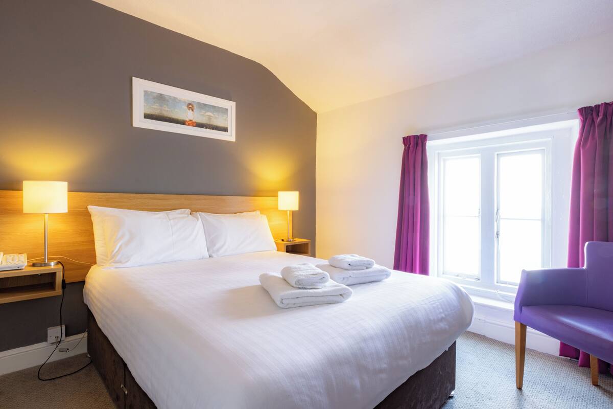 A hotel room with a large bed, two bedside lamps, a nightstand, a chair, and a window with curtains.