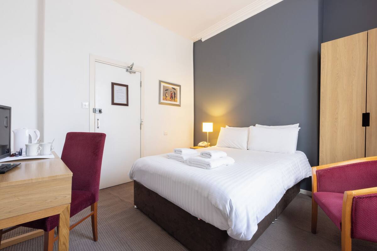 A hotel room with a bed, a desk, a chair, a wardrobe, and a nightstand with a lamp.