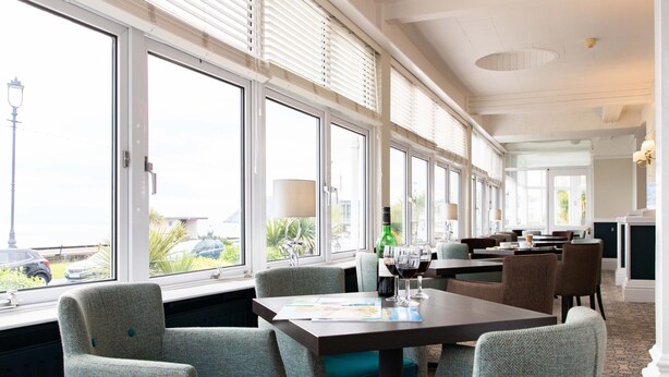 A hotel dining area with large windows, teal upholstered chairs, and a table with a menu and wine glasses. - The Marine Hotel (Llandudno)