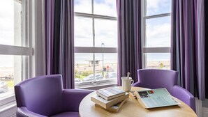 A room with purple chairs, a round table, and a window with a view of the beach and cars. - The Marine Hotel (Llandudno)