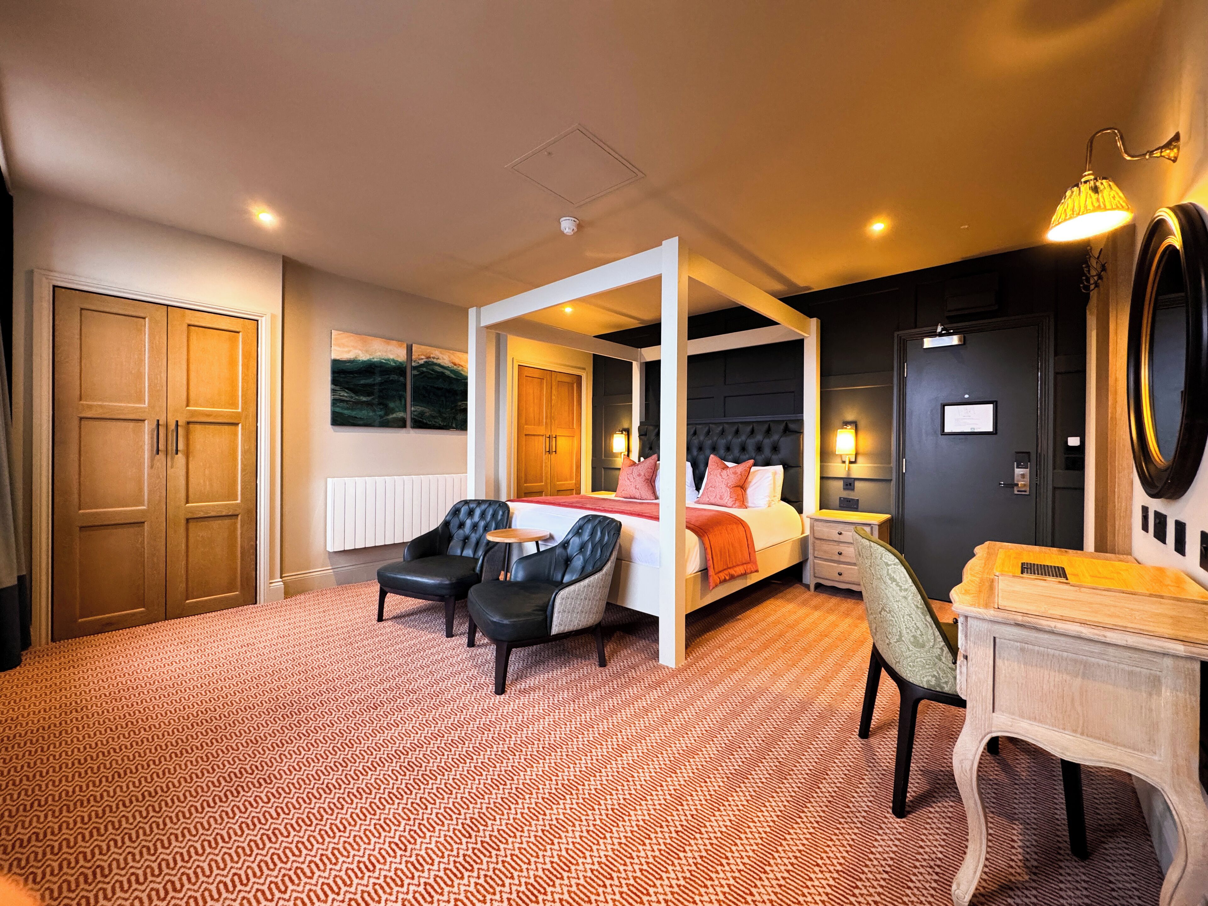 A four-poster bed with a white and orange bedspread, two black leather chairs, a wooden desk, and a large mirror.