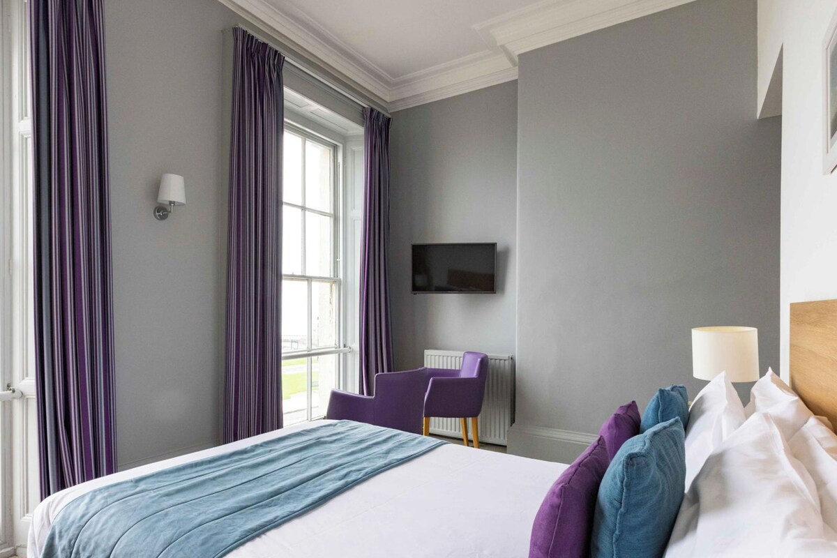 A hotel room with a large bed, purple curtains, a TV, and a window.