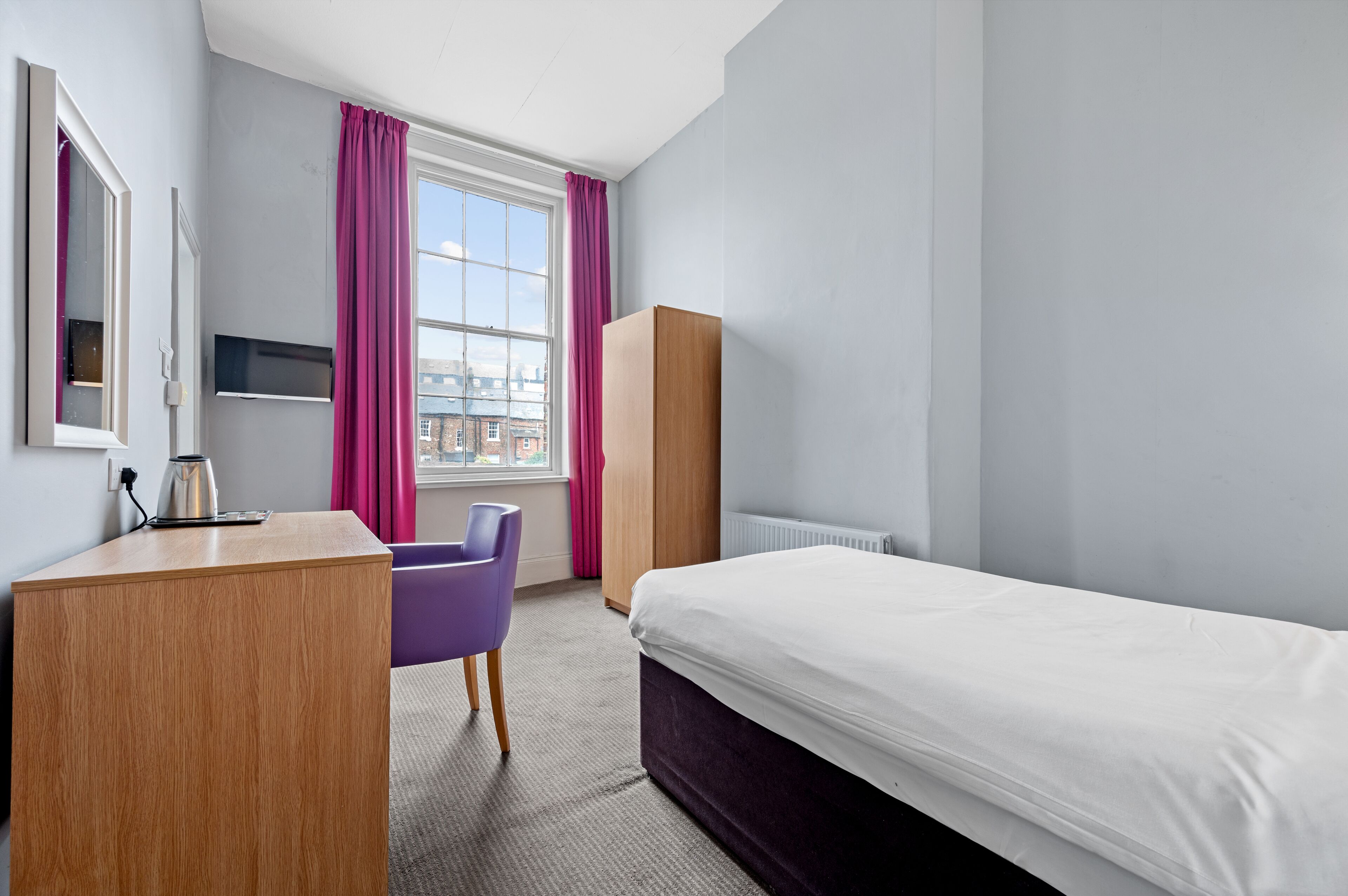 Hotellihuone, jossa on sänky, työpöytä, tuoli, vaatekaappi ja ikkuna, jossa on verhot.