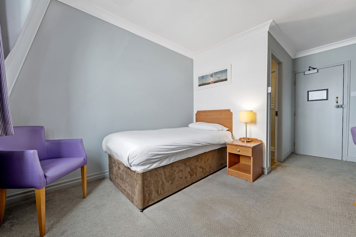 A bedroom with a bed, a nightstand with a lamp, a purple chair, and a door leading to another room.