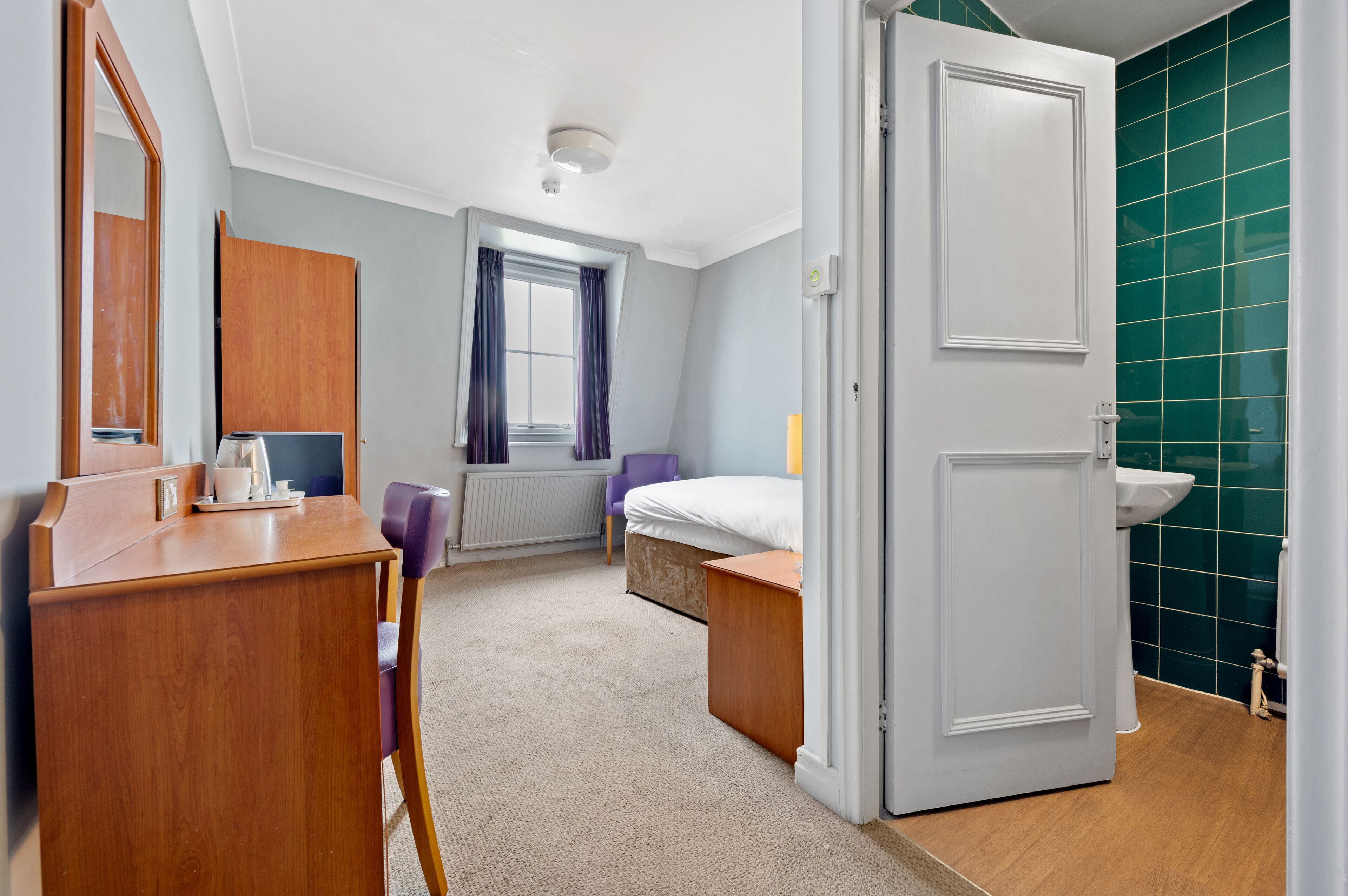 A hotel room with a wooden desk, a bed, a bathroom with a green tiled wall, and a window with purple curtains.