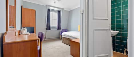 A hotel room with a wooden desk, a bed, a bathroom with a green tiled wall, and a window with purple curtains.