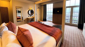 A hotel room with a large bed, a desk, a chair, a mirror, and a view of the city through large windows.